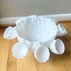 May include: A white milk glass punch bowl set featuring a large scalloped bowl with a fluted design and several matching small cups that hang from the rim of the bowl. The entire set is made of opaque white glass with a smooth, glossy finish.