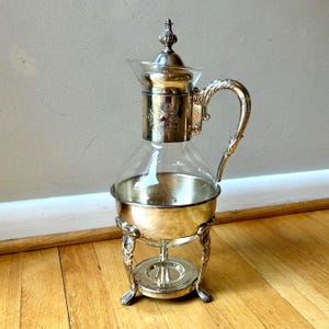 May include: A silver-plated glass carafe with a metal stand featuring three ornate legs. The carafe has a polished silver lid with a decorative finial, a clear glass body, and a silver base. The handle is curved and detailed with metal scrollwork.