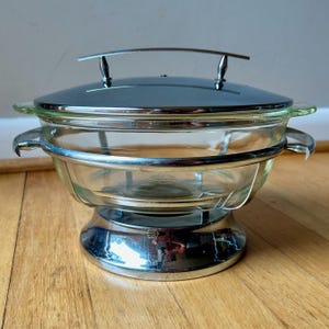 May include: A clear glass chafing dish with a chrome-plated metal frame and lid. The dish has a curved glass bowl, a lid with a handle, and side handles. The base is also chrome-plated. The chafing dish is designed for serving food.