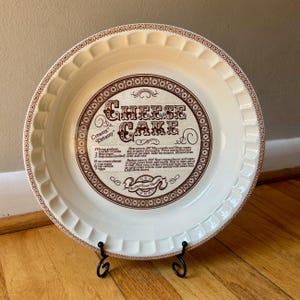 May include: A vintage white ceramic pie plate with a brown rim and a decorative border. The center features the words "CHEESE CAKE" in a stylized font, along with a recipe. The plate is displayed on a black metal stand.