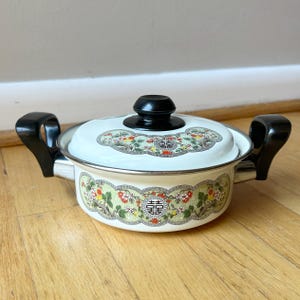 May include: A white enamel cooking pot with two black side handles and a matching lid featuring a black knob. The pot and lid are decorated with a green scalloped border, floral patterns in red and yellow, and a black double happiness symbol.