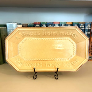May include: A rectangular, light yellow ceramic serving platter with the text "a joyous heart makes the loaves taste better" in embossed lettering. The platter is displayed on a black metal stand, with a rope-like border.