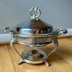 May include: A silver-colored chafing dish with a lid and ornate handles. The dish has a glass insert and sits on decorative legs. The lid has a decorative handle. The chafing dish is designed for serving food.