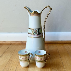 May include: A white porcelain pitcher with a gold handle and trim, accompanied by two matching cups. The pitcher and cups feature a decorative band with a pastoral scene and pink floral accents. The set is designed for serving beverages.