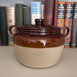 May include: A vintage, two-tone stoneware crock with a lid. The crock has a dark brown top and a lighter tan base. It features two handles and a knob on the lid. The background includes a collection of antique books.