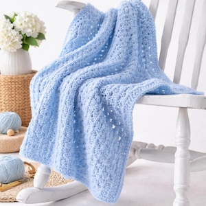 May include: A pale blue, hand-crocheted baby blanket draped over a white rocking chair. The blanket features a textured, open weave pattern. A white vase with white flowers and two balls of blue yarn are in the background.