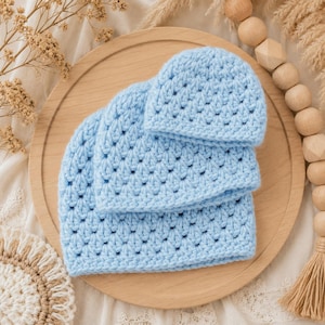 May include: A set of three light blue crochet hats stacked on top of each other. Each hat features a consistent textured stitch pattern throughout the body and a solid, smooth band along the bottom edge. The hats are crafted from soft yarn.