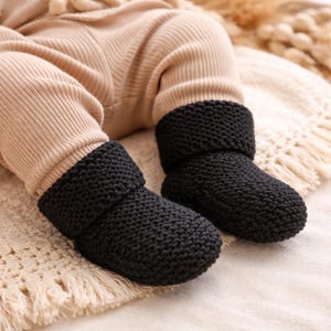 Puede incluir: Primer plano de los pies de un bebé con botines de punto negros. Los botines tienen un puño doblado y están hechos de un hilo grueso y texturizado. El bebé lleva pantalones acanalados beige. Los botines están sobre una manta color crema.