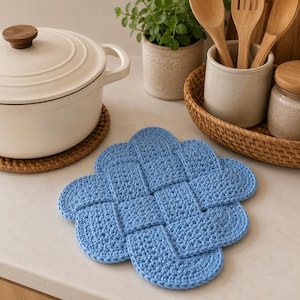 Può includere: Un sottopentola all'uncinetto color azzurro con un motivo intrecciato di forma quadrata e bordi arrotondati. L'accessorio tessile è realizzato in filato con una visibile trama a maglia, creando una superficie spessa per le stoviglie.