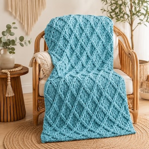 May include: A chunky knit throw blanket in a vibrant turquoise blue colour, featuring a prominent raised diamond pattern throughout the fabric. The blanket is draped over a light-coloured cushion on a curved wooden chair with a woven texture.