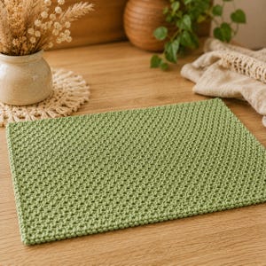 May include: A rectangular placemat in a solid sage green color, featuring a dense, uniform crochet stitch pattern with a textured surface. The placemat is constructed from thick yarn and displays a consistent, repeating knot-like weave throughout.