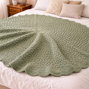 May include: A round, sage green crochet blanket with a scalloped edge is shown on a bed. The blanket has a textured pattern and is draped over white bedding. The bed is made up with pillows and a bedside table with a lamp.