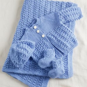 May include: A baby blue knitted set comprising a blanket, cardigan, hat, and booties. The set has a textured design, with white buttons on the cardigan. The blanket and hat are folded, and the booties are placed on the blanket.