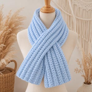 May include: A light blue scarf with a thick, ribbed, crocheted texture. The scarf is wrapped around a mannequin neck, featuring a consistent vertical stitch pattern throughout the length of the fabric, creating a soft and bulky appearance.