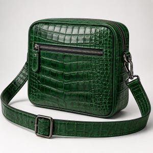 Puede incluir: Un bolso bandolera rectangular de color verde esmeralda intenso con una textura brillante de piel de cocodrilo. El bolso cuenta con un bolsillo frontal horizontal con cremallera, cierre superior de cremallera y una correa ajustable con hebilla metálica oscura.
