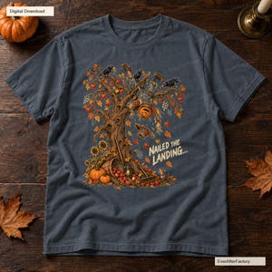 Pode incluir: Uma t-shirt cinzento-carvão com um gráfico de um espantalho com cabeça de abóbora pendurado de cabeça para baixo numa árvore. O design inclui folhas de outono, corvos, girassóis, abóboras e o texto "NAILED THE LANDING..." em letras brancas.