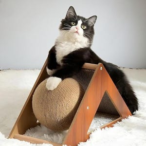 May include: A black and white cat lounges on a wooden cat scratcher with a sisal ball. The cat is positioned on top of the scratcher, with its paws resting on the ball. The scratcher has a triangular design.