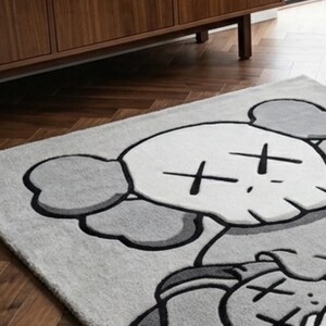 Pu&ograve; includere: Un tappeto rettangolare raffigurante un grande personaggio dei cartoni animati stilizzato con una testa simile a un teschio, orecchie rotonde e occhi a forma di X. Il design &egrave; realizzato in tonalit&agrave; grigio chiaro, grigio scuro e bianco.