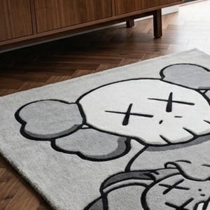 Pu&ograve; includere: Un tappeto rettangolare raffigurante un grande personaggio dei cartoni animati stilizzato con una testa simile a un teschio, orecchie rotonde e occhi a forma di X. Il design &egrave; realizzato in tonalit&agrave; grigio chiaro, grigio scuro e bianco.