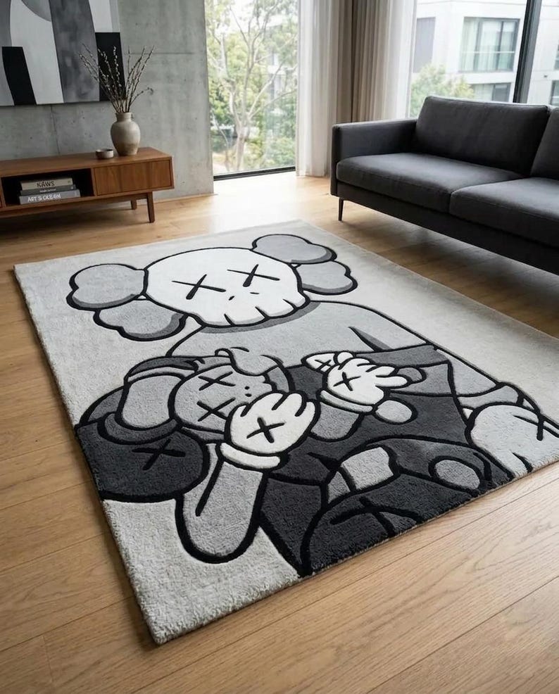 Pu&ograve; includere: Un tappeto rettangolare caratterizzato da un grande motivo grafico trapuntato nei toni del grigio, nero e bianco. Il disegno raffigura due personaggi dei cartoni animati stilizzati con occhi a forma di X, sovrapposti, in uno stile dai contorni marcati.