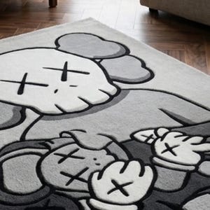 Pu&ograve; includere: Un tappeto rettangolare con una grande grafica di un personaggio dei cartoni animati stilizzato nei toni del grigio, bianco e nero. Il design raffigura due figure con occhi a forma di X e mani guantate, con una texture trapuntata su fondo grigio chiaro.