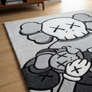 Pu&ograve; includere: Un tappeto rettangolare con una grande illustrazione grafica in grigio e bianco di un personaggio dei cartoni animati con occhi a forma di X e mani guantate. Il design include una testa simile a un teschio e una figura con le mani sul viso.