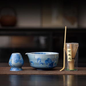 May include: A matcha tea set featuring a light blue ceramic bowl and a matching small cup, both adorned with a blue floral pattern. A bamboo whisk and scoop are included, along with a clear container holding the whisk. The set is arranged on a brown surface.