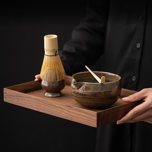 May include: A wooden serving tray holds a matcha tea set. The set includes a ceramic bowl, a bamboo whisk, and a wooden scoop. The bowl and whisk have brown and tan tones, and the tray is a dark wood.