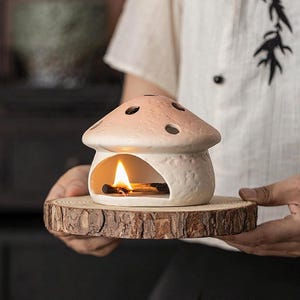 Puede incluir: Un calentador de aceite de cerámica con forma de hongo con una vela encendida en su interior. El calentador es de color rosa claro y blanco, con agujeros en la parte superior. Se encuentra sobre una rodaja de madera redonda y natural. La llama es visible a través de la abertura.