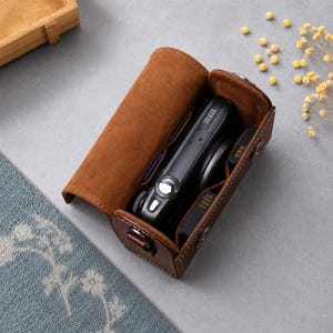 May include: A brown leather camera case with a flap lid, containing a black digital camera with a lens and a battery. The interior is lined with soft brown material, and the exterior features visible white stitching and metal snap fasteners.