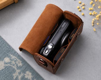 May include: A brown leather camera case with a flap lid, containing a black digital camera with a lens and a battery. The interior is lined with soft brown material, and the exterior features visible white stitching and metal snap fasteners.