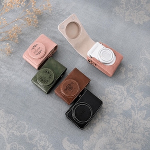 May include: Four rectangular camera cases in pink, olive green, brown, and black, each featuring a circular lens cover and custom engraving. One pink case is open, revealing a white compact camera inside with a circular lens and white body.