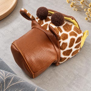 May include: A giraffe-themed pouch featuring a brown textured faux leather front section and a plush white and brown giraffe-patterned back. The pouch includes two dark brown pom-poms on top, small ears, and a bright yellow interior lining.