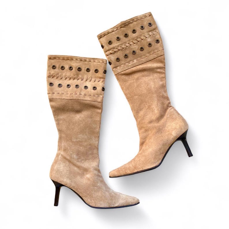 May include: Two tan suede knee-high boots with pointed toes and stiletto heels. The top of each boot features a decorative band with metal grommets and stitched details. The boots are set against a white background.