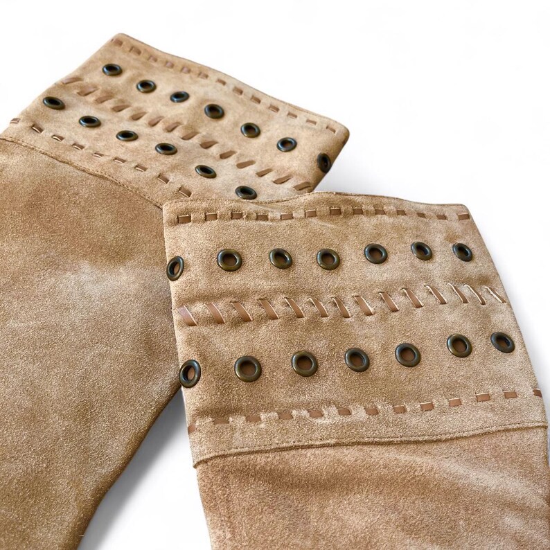 May include: Close-up of two tan suede boot cuffs. Each cuff features decorative stitching and metal grommets along the top edge. The cuffs are designed to be worn over boots, adding a stylish layer.