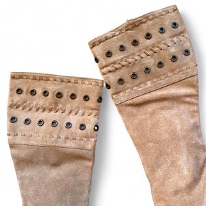May include: Tan suede boots with decorative metal grommets and stitched detailing. The boots have a knee-high design and a soft, textured appearance. The upper portion of the boots features a series of evenly spaced grommets and decorative stitching.