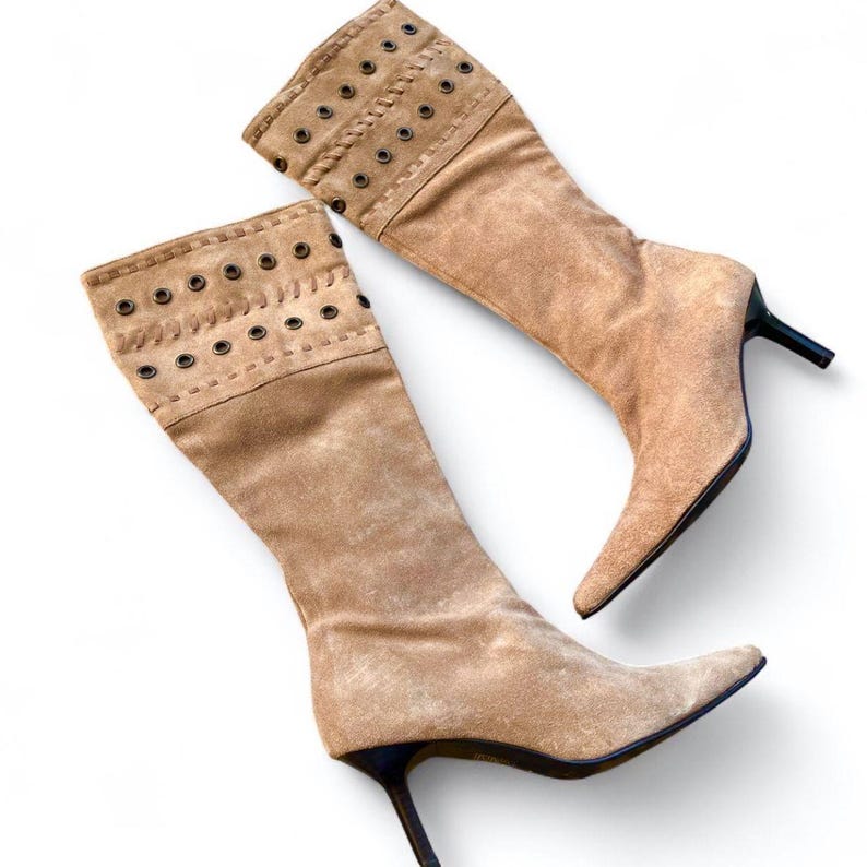 May include: A pair of tan suede knee-high boots with pointed toes and stiletto heels. The upper cuff features decorative grommets and stitching. The boots are set against a white background.