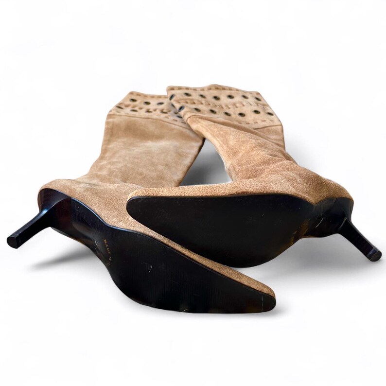 May include: A pair of tan suede boots with pointed toes and stiletto heels. The boots have a perforated design at the top and dark brown soles. The heels are approximately 3 inches tall.