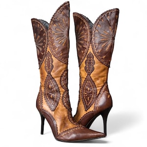 以下が含まれることがあります： 茶色とタン色の革ブーツのペア。複雑な切り抜きとステッチのデザインが施されています。ブーツはポインテッドトゥで、細い黒いハイヒールが付いています。上部は装飾的なスカラップエッジが特徴です。
