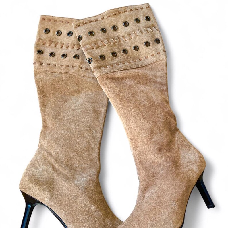 May include: A pair of knee-high tan suede boots with black stiletto heels. The boots have a folded-over cuff with decorative grommets and stitching. The boots are made of a soft, textured material.