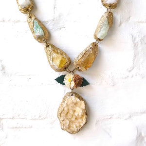 May include: A gold necklace with a large, oval-shaped crystal pendant. The pendant is made of a clear, white crystal with a gold-painted exterior. The necklace is made of smaller, gold-painted crystals in various shapes and sizes. The necklace is accented with a small, green leaf and a cluster of white and brown flowers.