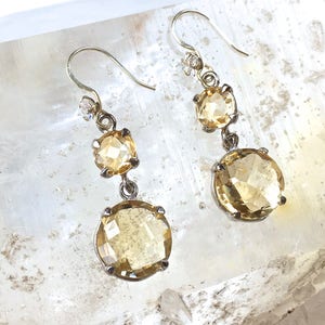 May include: A pair of gold tone dangle earrings with three faceted citrine stones. The stones are round and have a light yellow color.