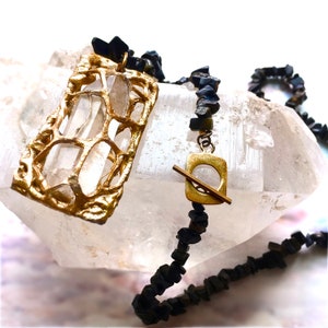 Puede incluir: Un collar de oro con un colgante geométrico y cuentas de piedras preciosas negras. El collar se muestra en un cristal de cuarzo transparente.