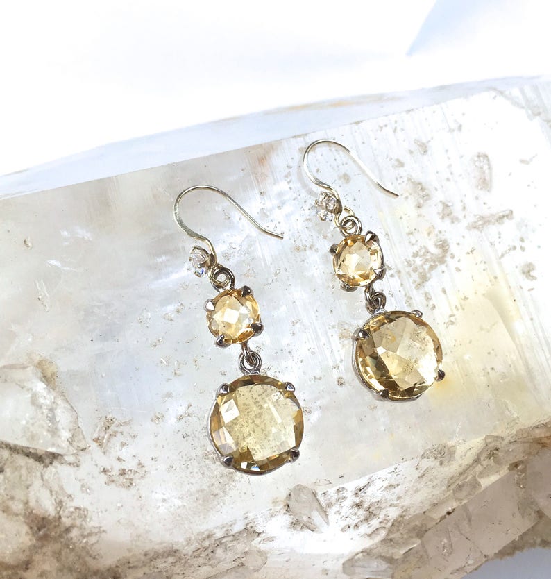 May include: A pair of gold tone dangle earrings with three faceted citrine stones. The stones are round and have a light yellow color.