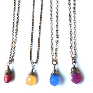 May include: Four gold-toned necklaces with teardrop-shaped pendants in red, yellow, blue, and purple. Each pendant is wire-wrapped and hangs from a delicate chain. The pendants have a faceted, gemstone appearance.