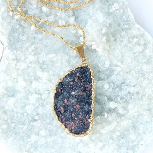 May include: A gold chain necklace with a large, dark gray and copper-colored stone pendant. The pendant is shaped like a teardrop and has a rough, natural texture.