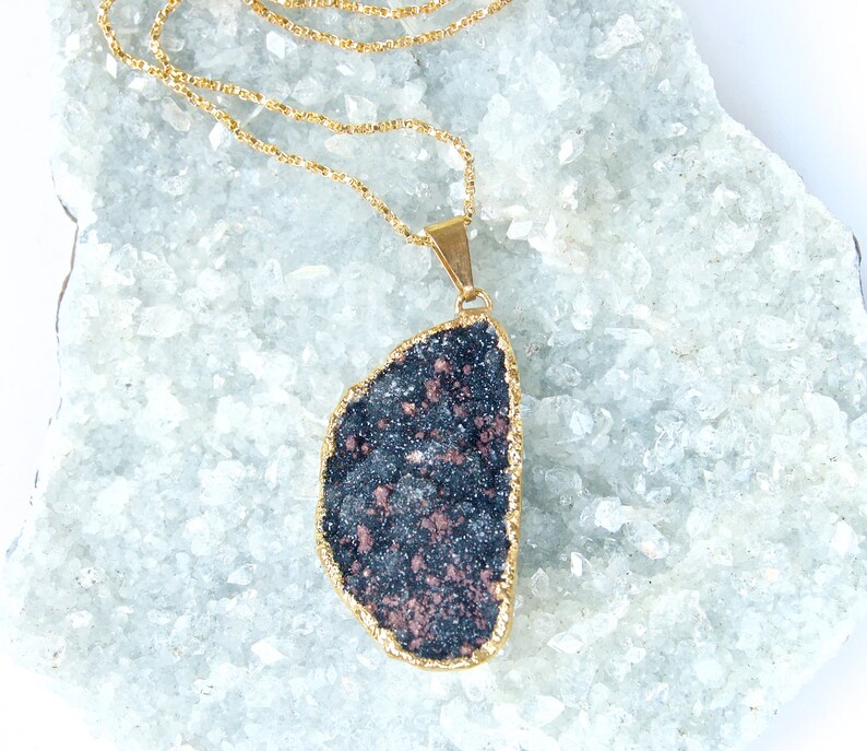 May include: A gold chain necklace with a large, dark gray and copper-colored stone pendant. The pendant is shaped like a teardrop and has a rough, natural texture.