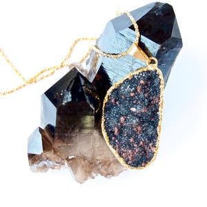 May include: A gold chain necklace with a large, dark grey and copper-colored gemstone pendant. The pendant is shaped like a teardrop and is set against a cluster of clear quartz crystals.