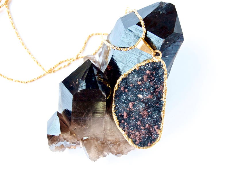 May include: A gold chain necklace with a large, dark grey and copper-colored gemstone pendant. The pendant is shaped like a teardrop and is set against a cluster of clear quartz crystals.
