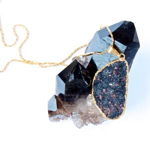 May include: A gold chain necklace with a large, dark gray, irregularly shaped pendant. The pendant is surrounded by clear and black quartz crystals.